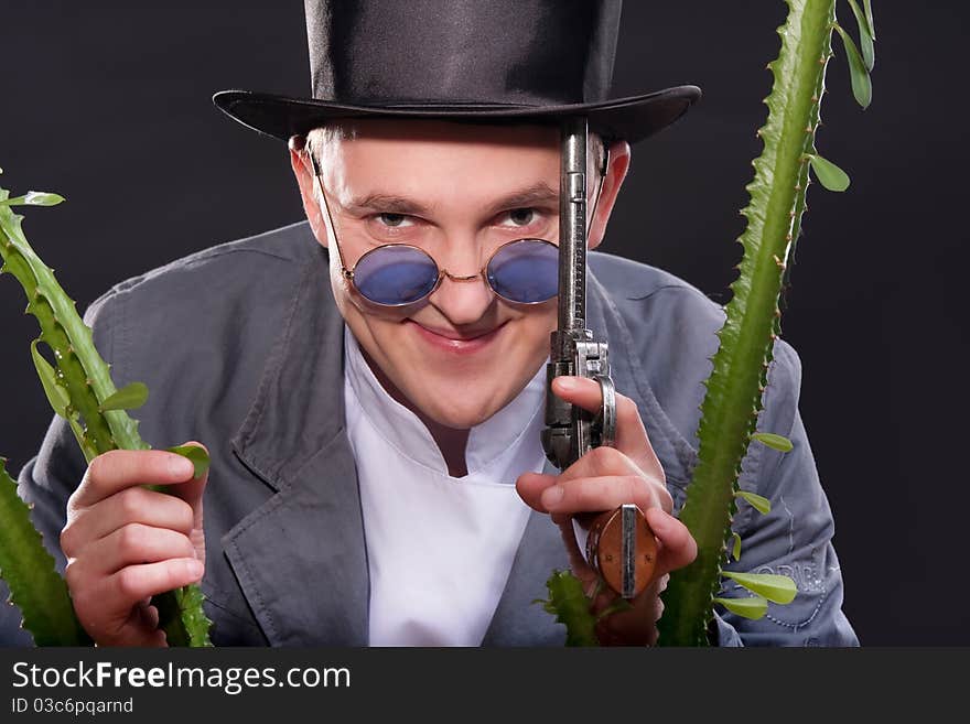 Emotional portrait of a man in a hat with a gun in his hand peek out from behind a plant