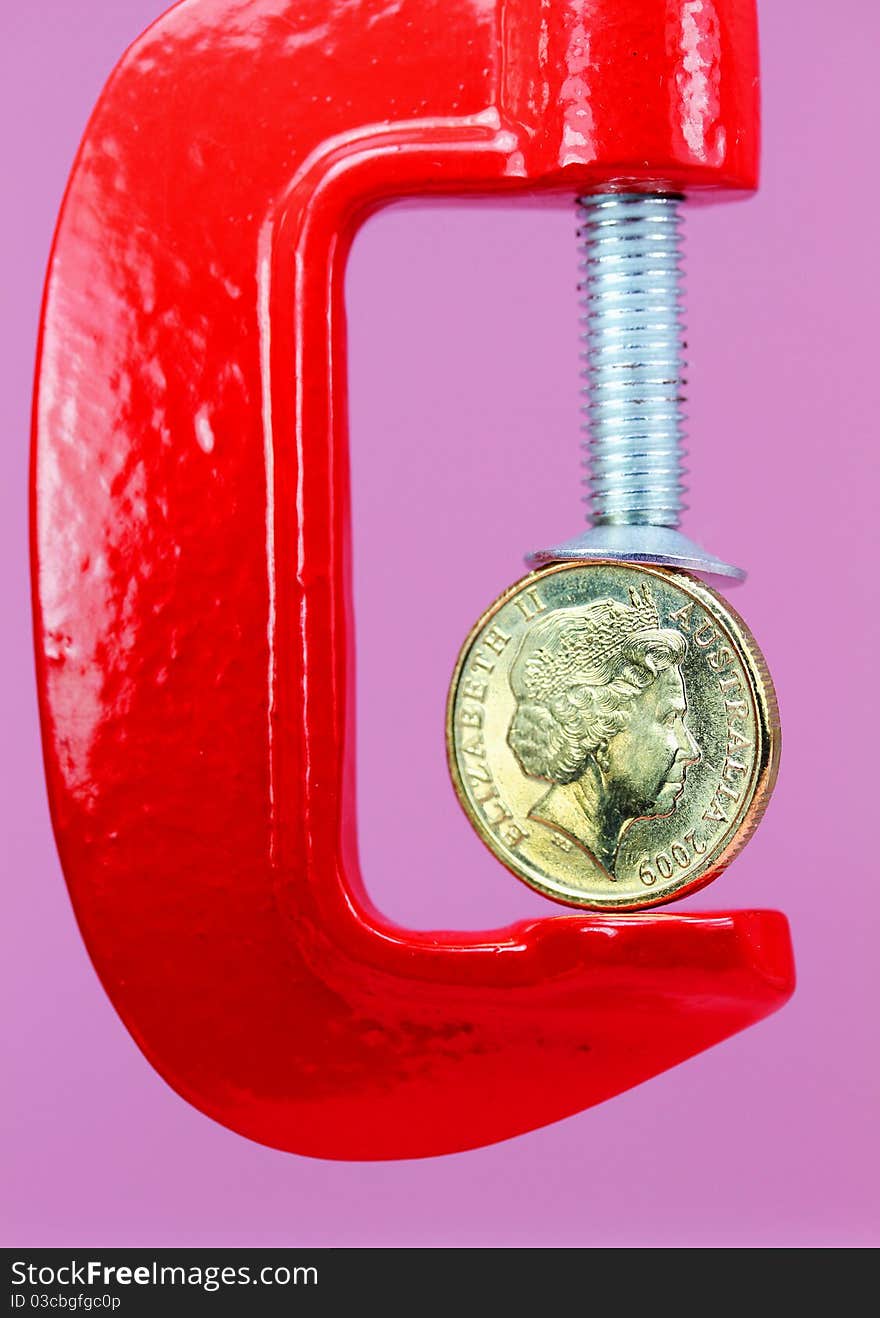 A gold coin held in a red clamp with a light pastel green background, indicating the pressure is on currency during these uncertain times. A gold coin held in a red clamp with a light pastel green background, indicating the pressure is on currency during these uncertain times.