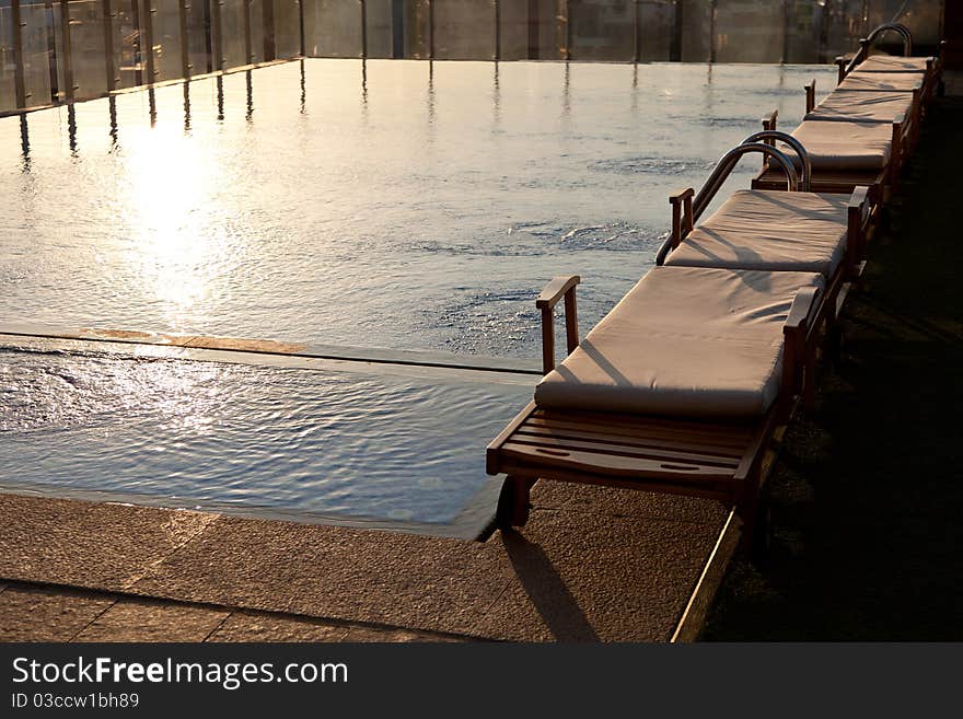 A row of cushioned benches by the side of a hotel pool at sunset. A row of cushioned benches by the side of a hotel pool at sunset.