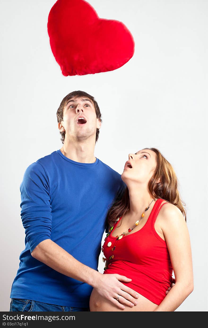 Surprised couple of young man and pregnant woman looking up at red heart