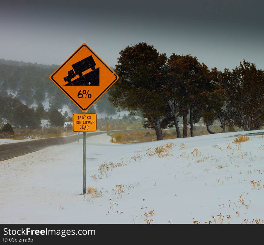 A photograph of a 6% Grade Sign. The sign states, Trucks Use Lower Gear. It is a snowy icy day on a windy road. A photograph of a 6% Grade Sign. The sign states, Trucks Use Lower Gear. It is a snowy icy day on a windy road.