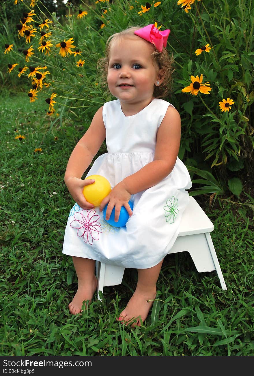 Beautiful blonde headed girl holding balls looking at the camera smiling while sitting on a stool outside on a sunny day. Beautiful blonde headed girl holding balls looking at the camera smiling while sitting on a stool outside on a sunny day