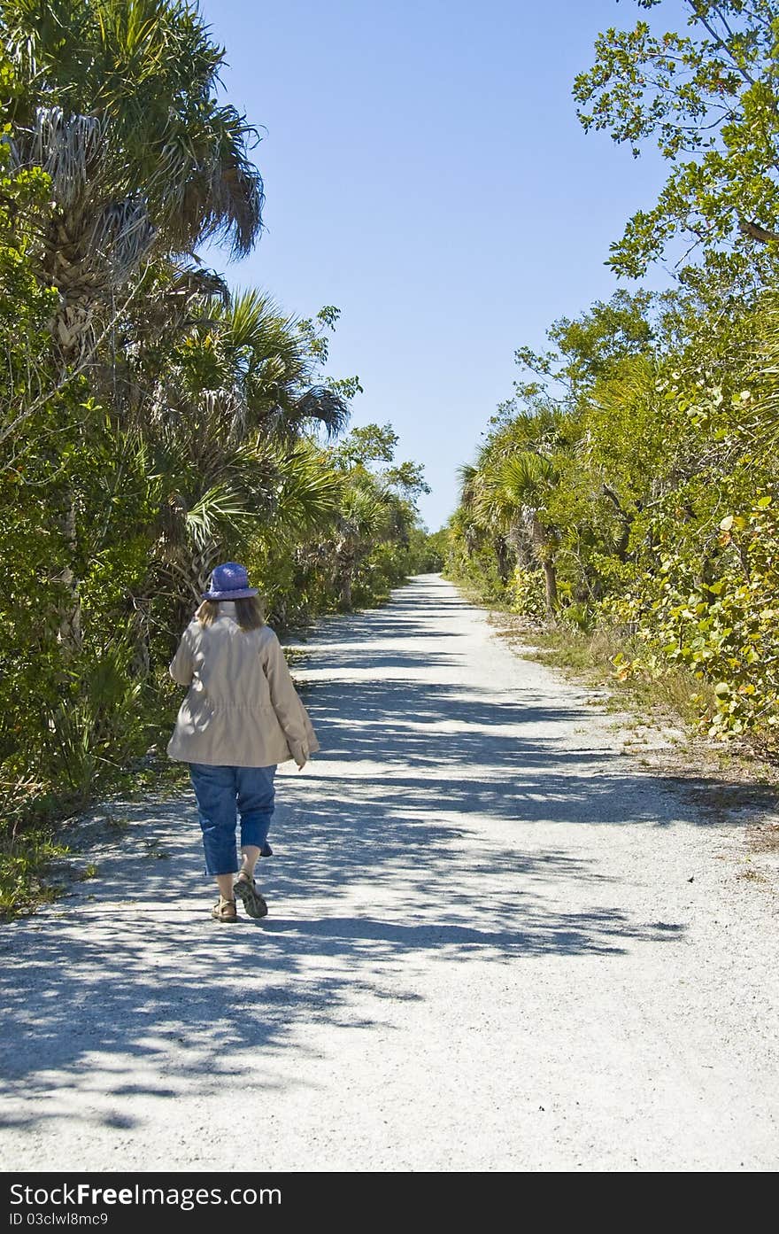 Woman walking down a sandy path surrounded on both sides by green foliage. View from behind. Path leads straight away from camera. Woman walking down a sandy path surrounded on both sides by green foliage. View from behind. Path leads straight away from camera.