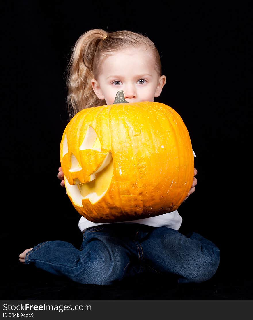 A young child is holding a pumpkin. The pumpkin is sideways and the child is kneeling. A young child is holding a pumpkin. The pumpkin is sideways and the child is kneeling.