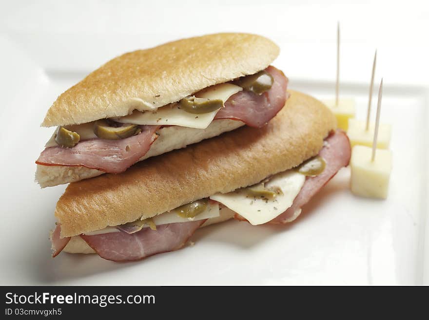 White bread sandwiches filled with smoked ham green olives and sliced cheese on white square plate accompanied with cheese cubes. White bread sandwiches filled with smoked ham green olives and sliced cheese on white square plate accompanied with cheese cubes