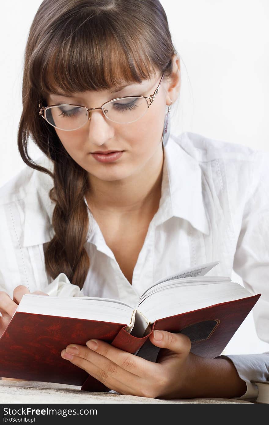 Beautiful girl with glasses in a white shirt thoughtfully reading a book. Her Hair is braided into a braid. Focus on the forefront of the book. Close-up. Photographed in a studio against a white background. Beautiful girl with glasses in a white shirt thoughtfully reading a book. Her Hair is braided into a braid. Focus on the forefront of the book. Close-up. Photographed in a studio against a white background.