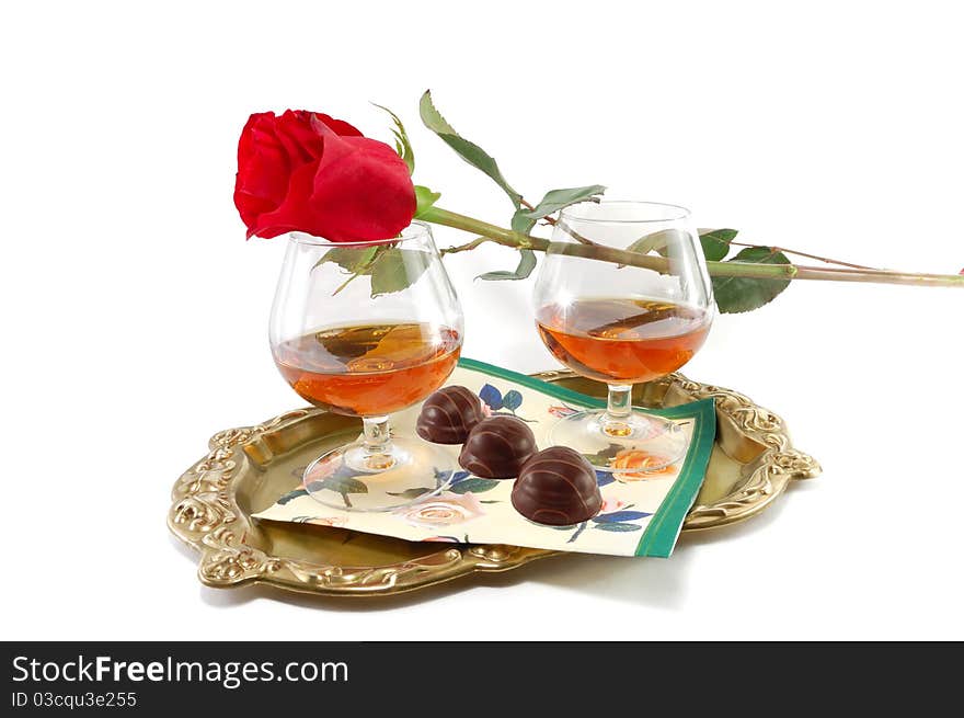 The rose and glasses with cognac and sweets on a tray are isolated on a white background