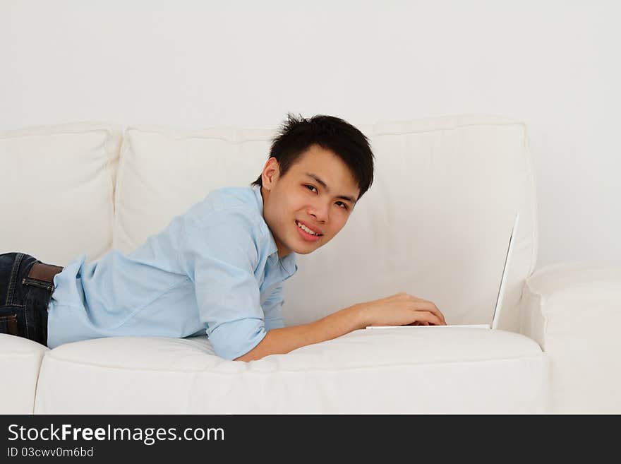 A smiling young man lying prone while using his laptop on a sofa. A smiling young man lying prone while using his laptop on a sofa