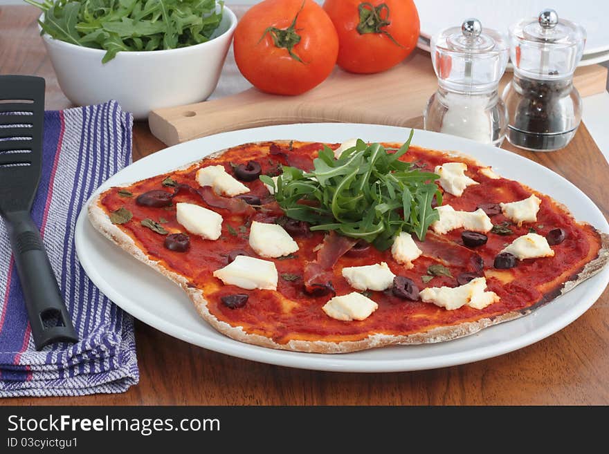 A delicious homemade pizza topped with prosciutto, ricotta cheese, olives and rocket salad. On a wooden kitchen table ready to be served. A delicious homemade pizza topped with prosciutto, ricotta cheese, olives and rocket salad. On a wooden kitchen table ready to be served.