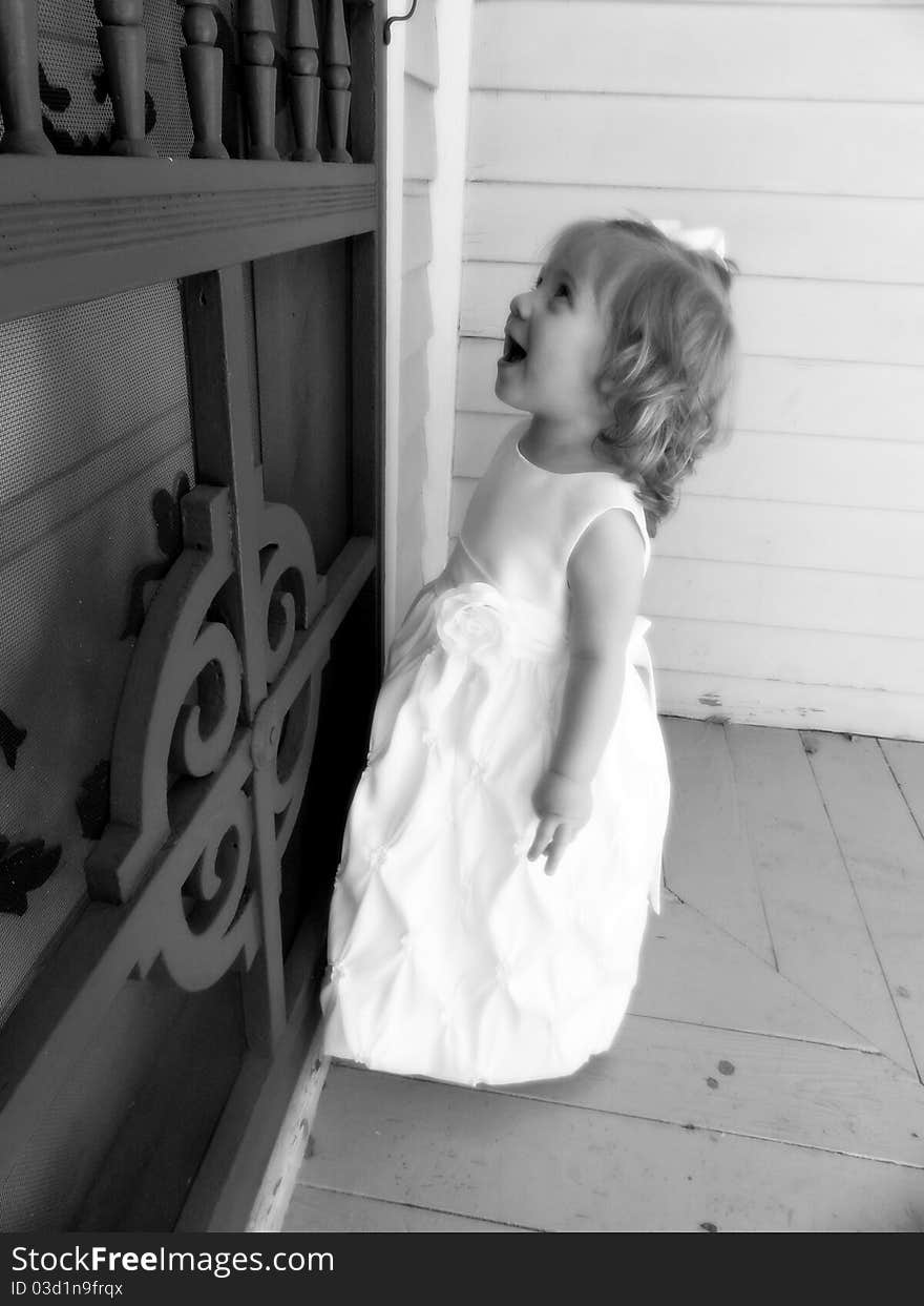 A baby girl is happy while looking at designes on screen door of an old colonial house. She is wearing a white formal dress and white bow. A baby girl is happy while looking at designes on screen door of an old colonial house. She is wearing a white formal dress and white bow.