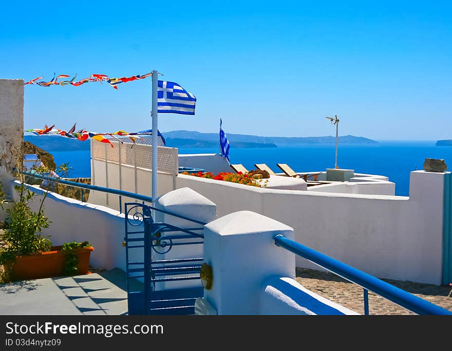 Beautiful white terrace at resort on island of Santorini in Oia, Greece. Traditional greek architecture and national flag of Greece. Aegean sea in a background. Beautiful white terrace at resort on island of Santorini in Oia, Greece. Traditional greek architecture and national flag of Greece. Aegean sea in a background.