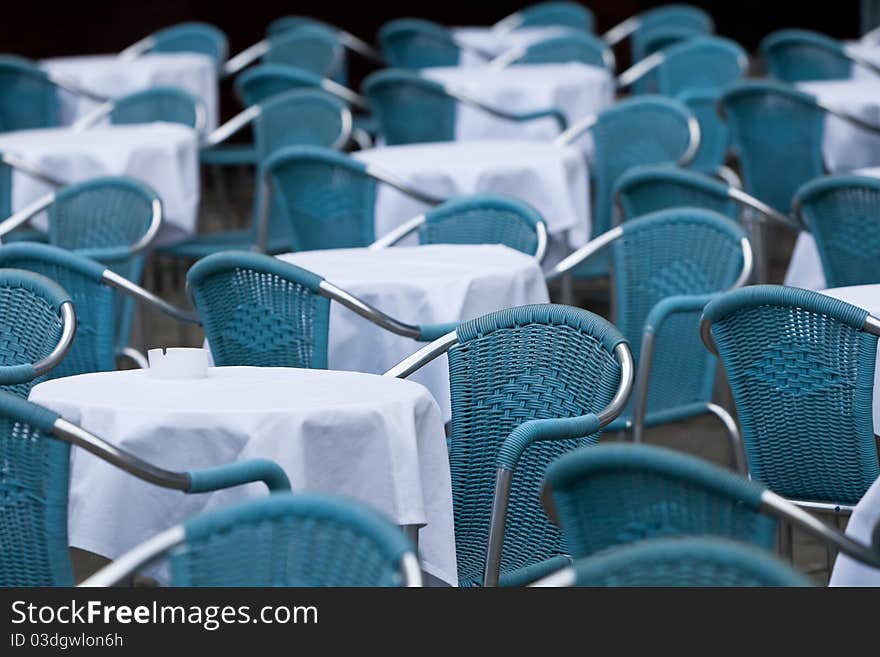 Many empty blue chairs with white rounded tables outside in front of restaurant. Many empty blue chairs with white rounded tables outside in front of restaurant.