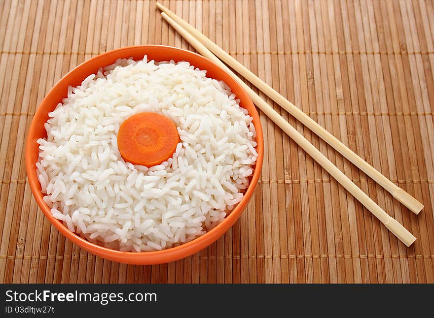 Orange bowl of cooked rice decorated with carrots and sticks on bamboo napkin. Orange bowl of cooked rice decorated with carrots and sticks on bamboo napkin.