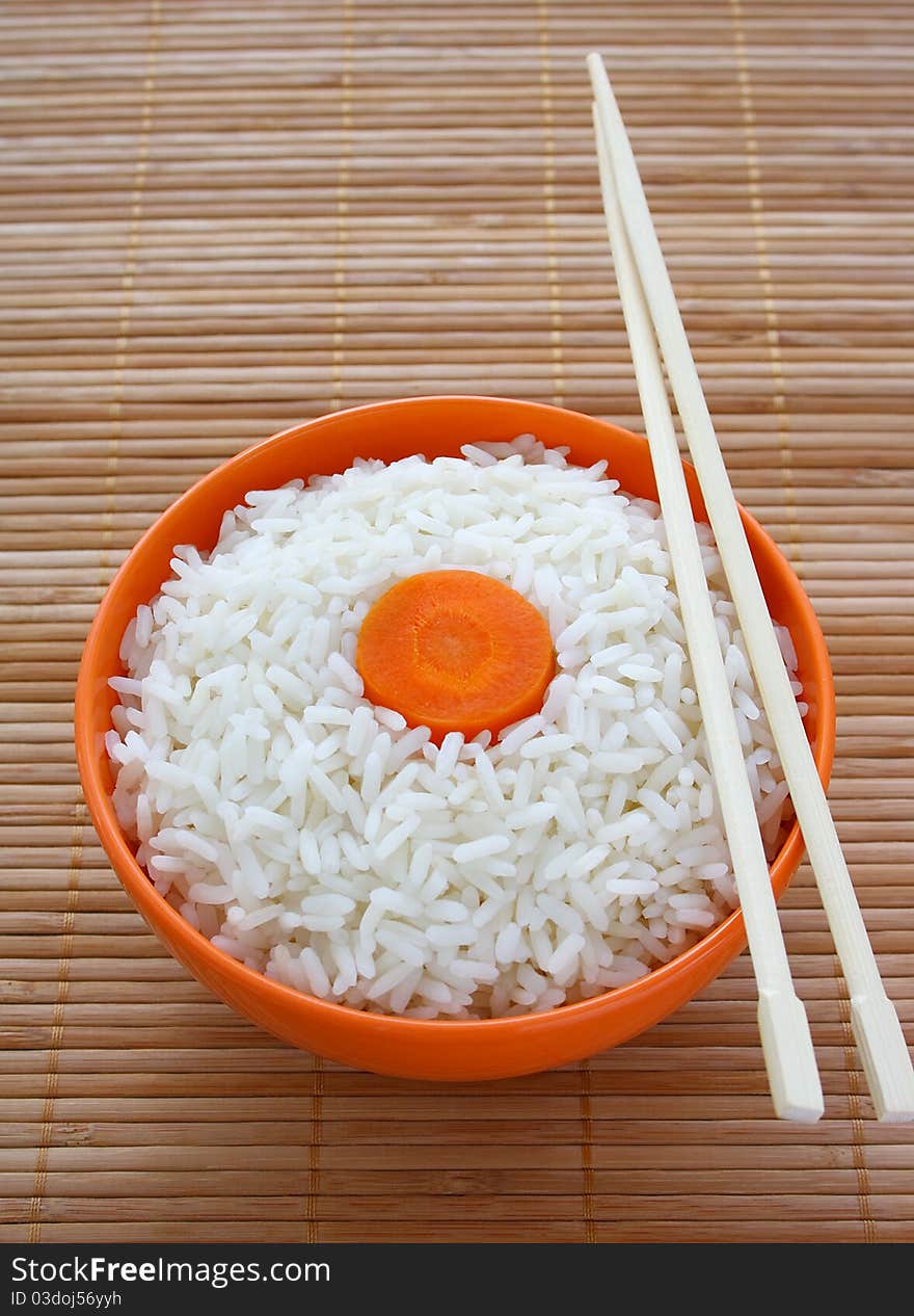 Orange bowl of cooked rice decorated with carrots and sticks on bamboo napkin. Orange bowl of cooked rice decorated with carrots and sticks on bamboo napkin.