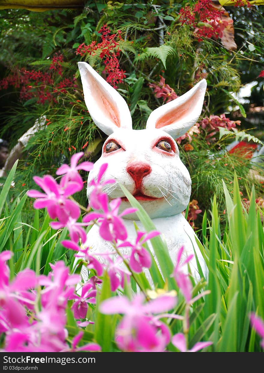 A statue of a white, pink-nosed rabbit sitting in a garden surrounded by flowers. A statue of a white, pink-nosed rabbit sitting in a garden surrounded by flowers