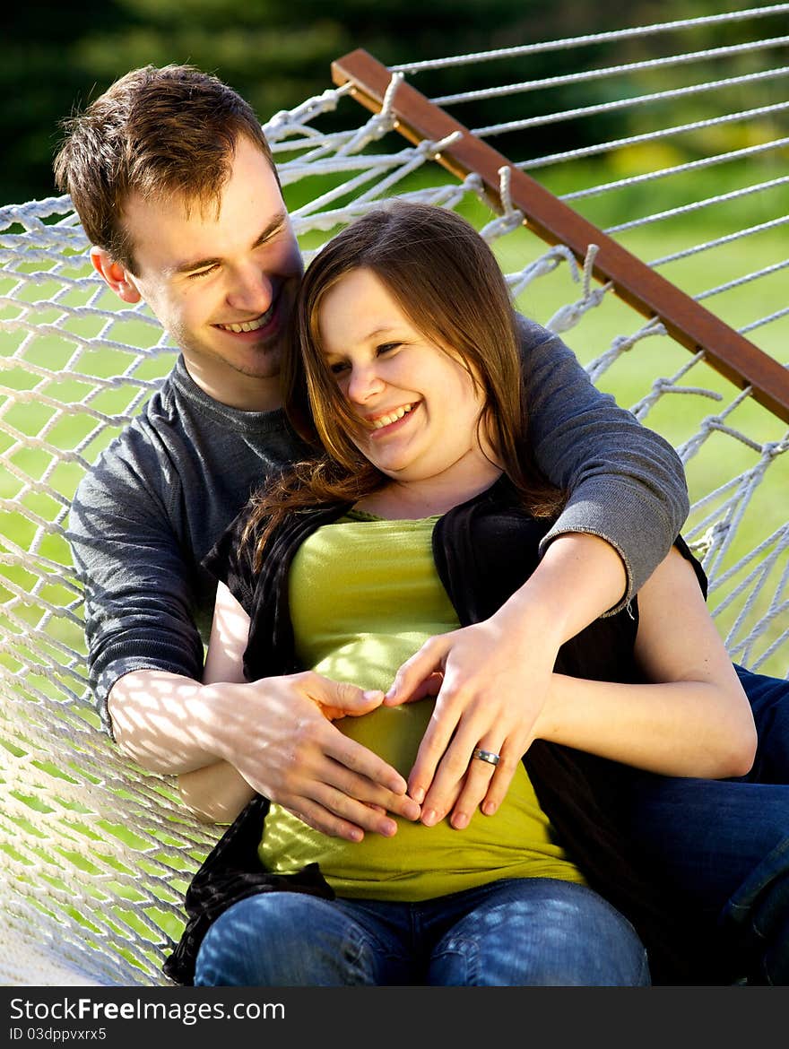 Young couple sitting on a hammock laughing with their hands on her pregnant belly. Young couple sitting on a hammock laughing with their hands on her pregnant belly.