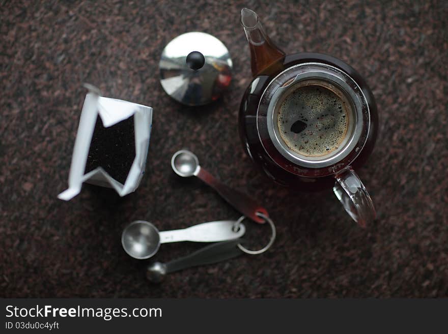 Bag of tea leaves, measuring spoons and teapot full of water and tea on a granite countertop, viewed from above. Bag of tea leaves, measuring spoons and teapot full of water and tea on a granite countertop, viewed from above