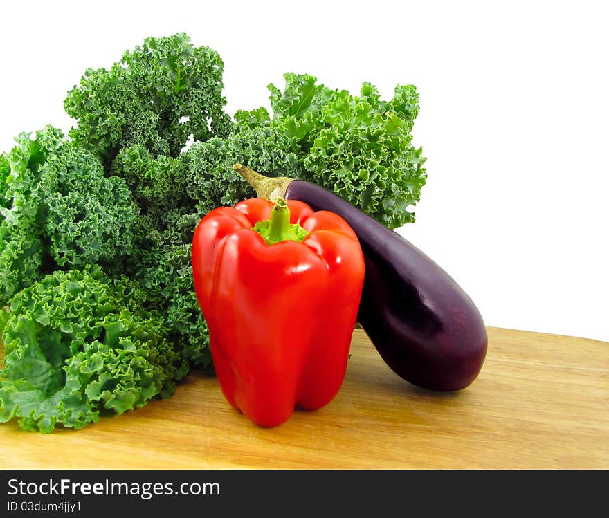 A red bell pepper, a purple eggplant, and a bunch of green kale on a wooden cutting board isolated on white. A red bell pepper, a purple eggplant, and a bunch of green kale on a wooden cutting board isolated on white.