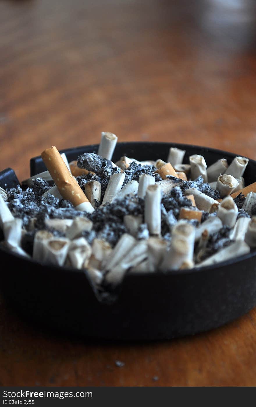 Close up of a black ash tray filled with stubbed out cigarettes on a brown wooden table. Close up of a black ash tray filled with stubbed out cigarettes on a brown wooden table