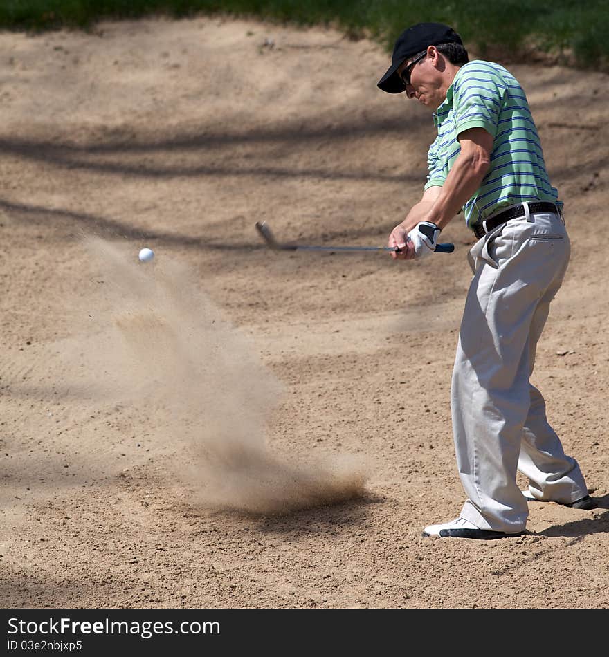 Golfer hits his golf ball out of the sand trap with the call frozen in the image after being hit.