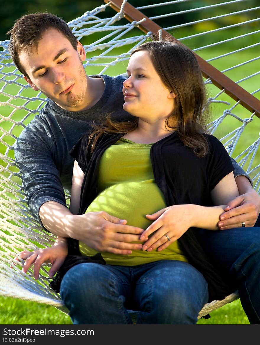 Young man sits on a hammock admiring his wife’s pregnant belly by making a heart with his hands. Young man sits on a hammock admiring his wife’s pregnant belly by making a heart with his hands.