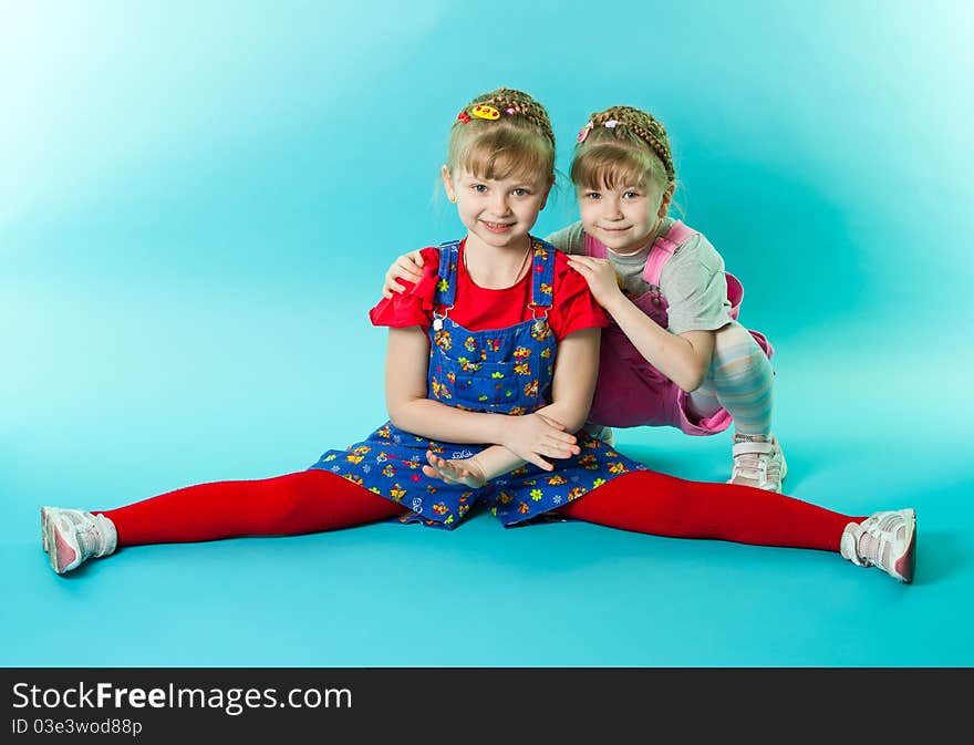 A girl trying to do the splits exercise, her twin sister sitting besides her over blue background. A girl trying to do the splits exercise, her twin sister sitting besides her over blue background