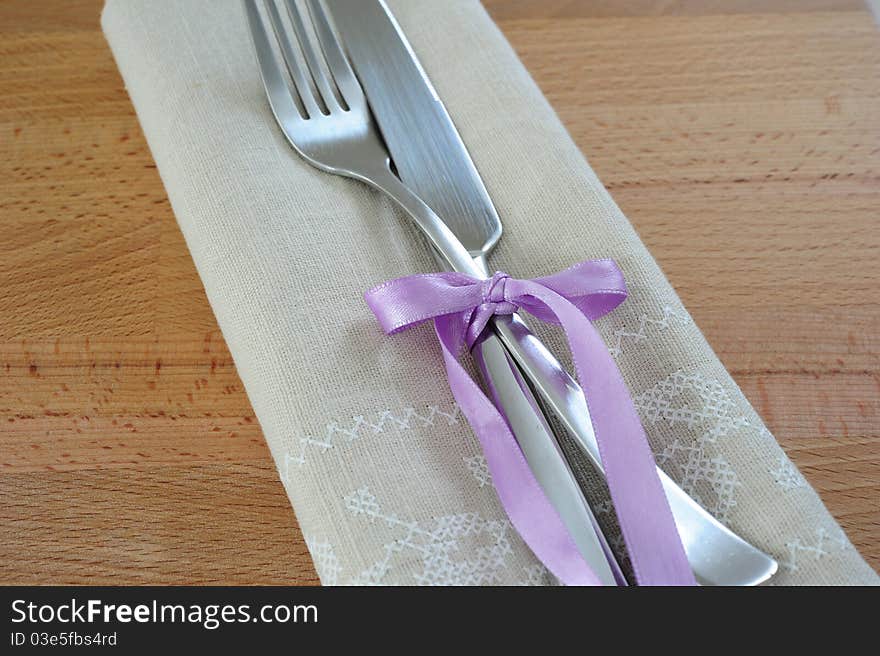 Fork and knife, tied with purple ribbon, with a linen cloth on a wooden table. Fork and knife, tied with purple ribbon, with a linen cloth on a wooden table