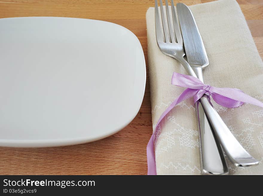 White plate, fork and knife, tied with purple ribbon, with a linen cloth on a wooden table. White plate, fork and knife, tied with purple ribbon, with a linen cloth on a wooden table