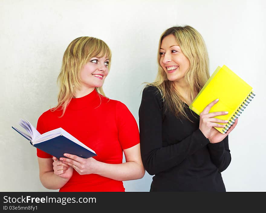 Two beautiful young girl with long blond hair are holding a book and notebook, look at each other, smiling. Two beautiful young girl with long blond hair are holding a book and notebook, look at each other, smiling