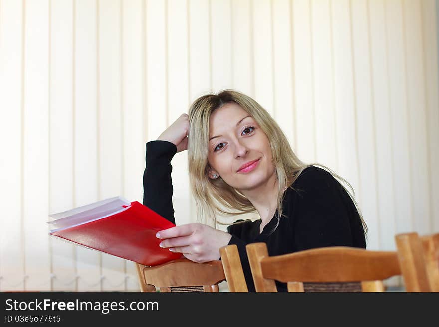Beautiful young girl with long blond hair and dark eyes sits leaning back in his chair with a red folder in his hands. Beautiful young girl with long blond hair and dark eyes sits leaning back in his chair with a red folder in his hands