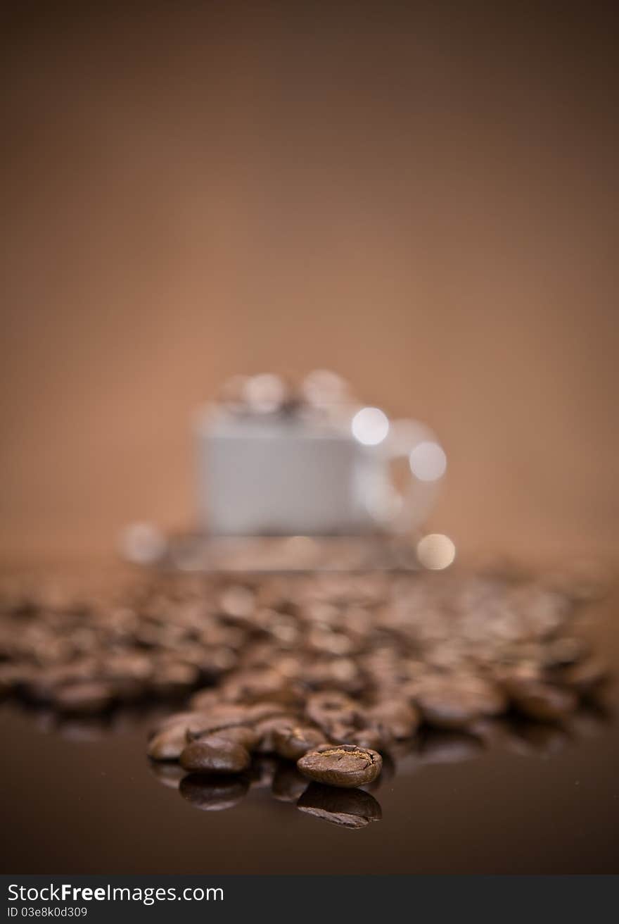 Studio macro shot beans of coffee on black miror with veri blurred white ceramic coffee cup on brown background with copy space selective focus. Studio macro shot beans of coffee on black miror with veri blurred white ceramic coffee cup on brown background with copy space selective focus