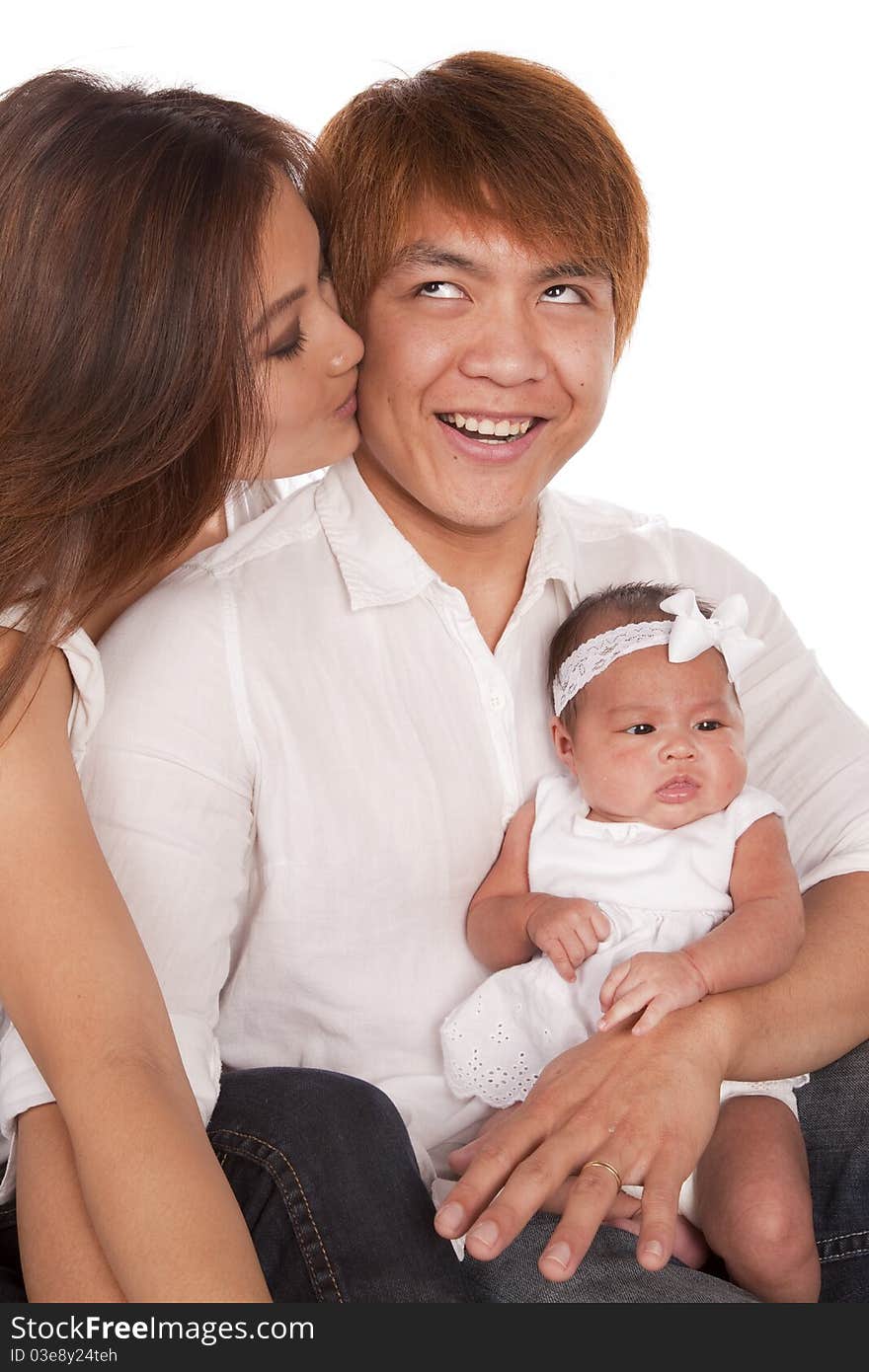 A mother kissing her husband on the cheek while he is holding their baby girl. The father has a funny expression on his face. A mother kissing her husband on the cheek while he is holding their baby girl. The father has a funny expression on his face.