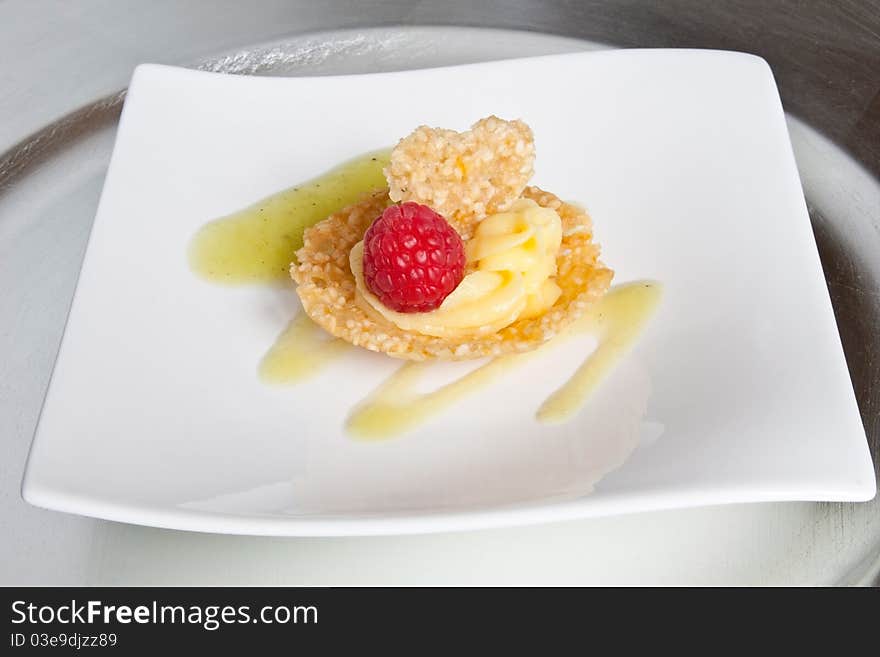 A dessert consisting of a sesame seed tuille set on kiwi coulis, filled with passionfruit fondant & garnished with a raspberry and heart-shaped tuille. Served on a white plate on a silver charger. A dessert consisting of a sesame seed tuille set on kiwi coulis, filled with passionfruit fondant & garnished with a raspberry and heart-shaped tuille. Served on a white plate on a silver charger.