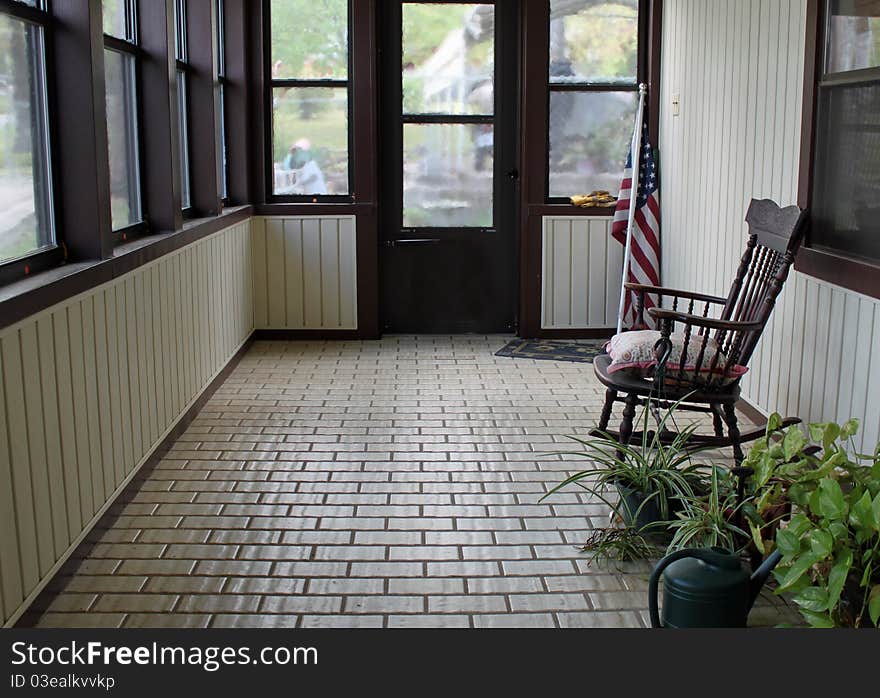 Enclosed, tiled, porch with potted plants, a watering can, an antique rocker and an American Flag. Enclosed, tiled, porch with potted plants, a watering can, an antique rocker and an American Flag.