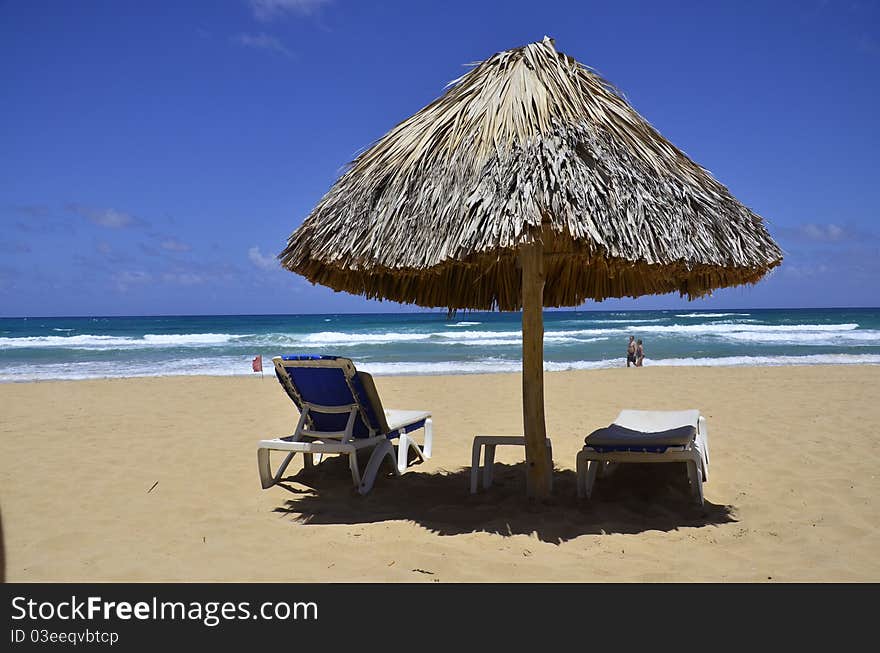 A palm frond umbrella shades 2 lounge chairs at the beach. A palm frond umbrella shades 2 lounge chairs at the beach