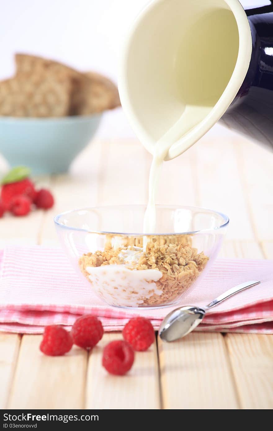 Pouring milk on a bowl of cereal (muesli/granola) with fresh strawberries, raspberries and fruit in the background. Pouring milk on a bowl of cereal (muesli/granola) with fresh strawberries, raspberries and fruit in the background.