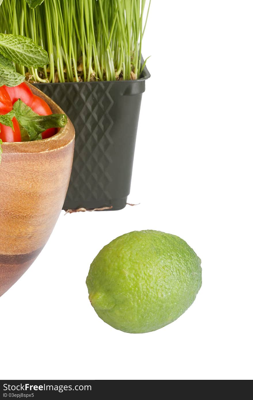 Green lime isolated on a white background next to wheatgrass and a wooden plate with red bell peppers.