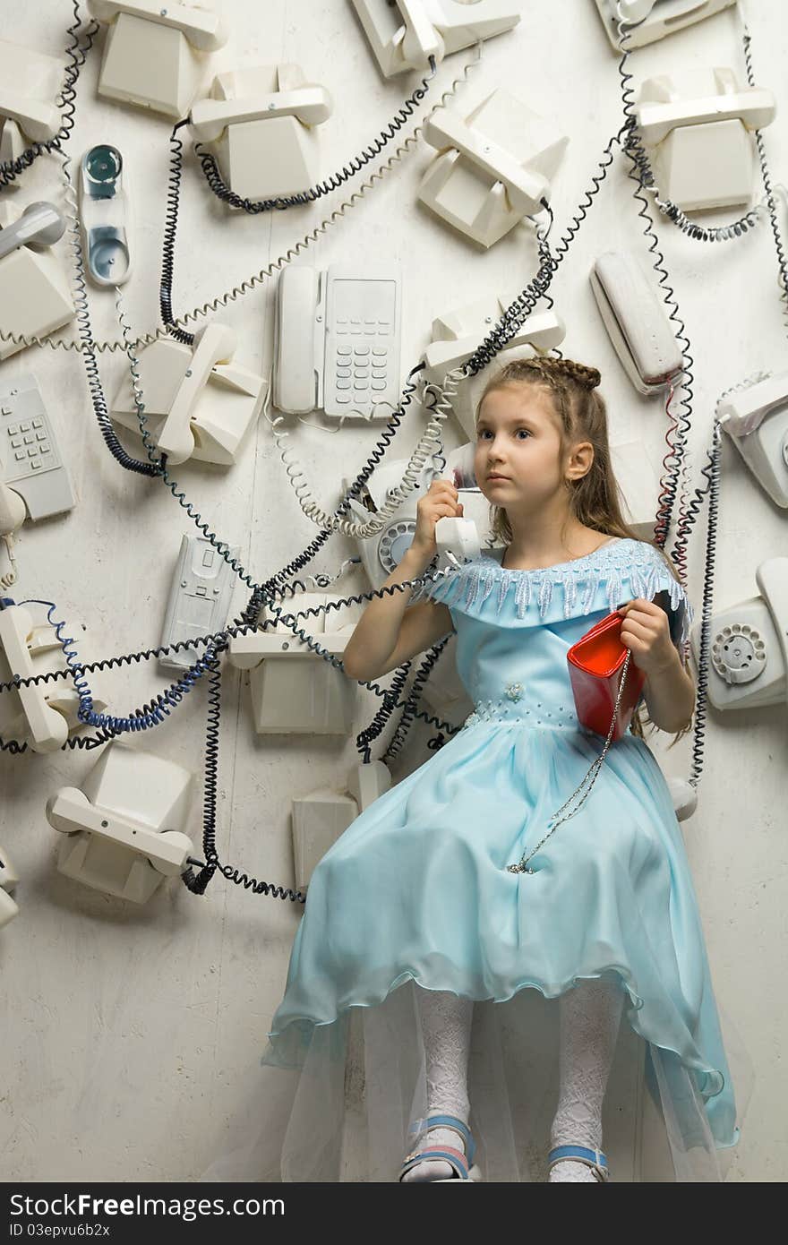 Image of a beautiful seriously little girl sitting on the white cube and holding a phone surrounded by many phones on the wall. Image of a beautiful seriously little girl sitting on the white cube and holding a phone surrounded by many phones on the wall