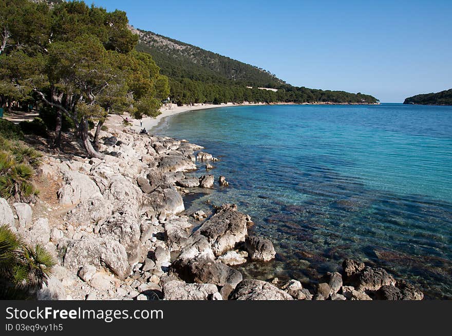 Image shows coast of Formentor, Majorca, Spain. Stones an trees on the left, blue sea on the right, blue sky above.