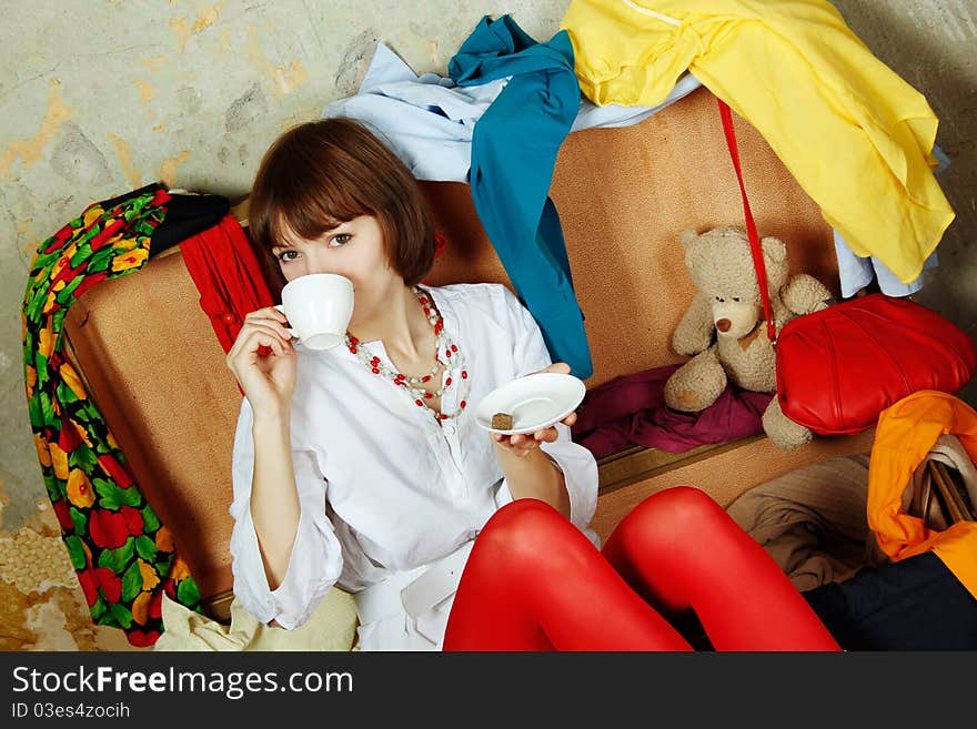 Young beautiful woman on the background of a concrete wall system sits in a big old-fashioned suitcase filled with clothes. Drinking coffee. Young beautiful woman on the background of a concrete wall system sits in a big old-fashioned suitcase filled with clothes. Drinking coffee