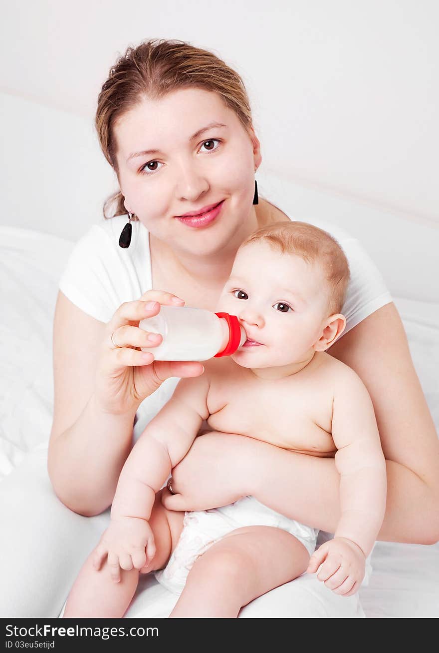 Young mother feeding her her six months old baby with milk  on the bed at home. Young mother feeding her her six months old baby with milk  on the bed at home