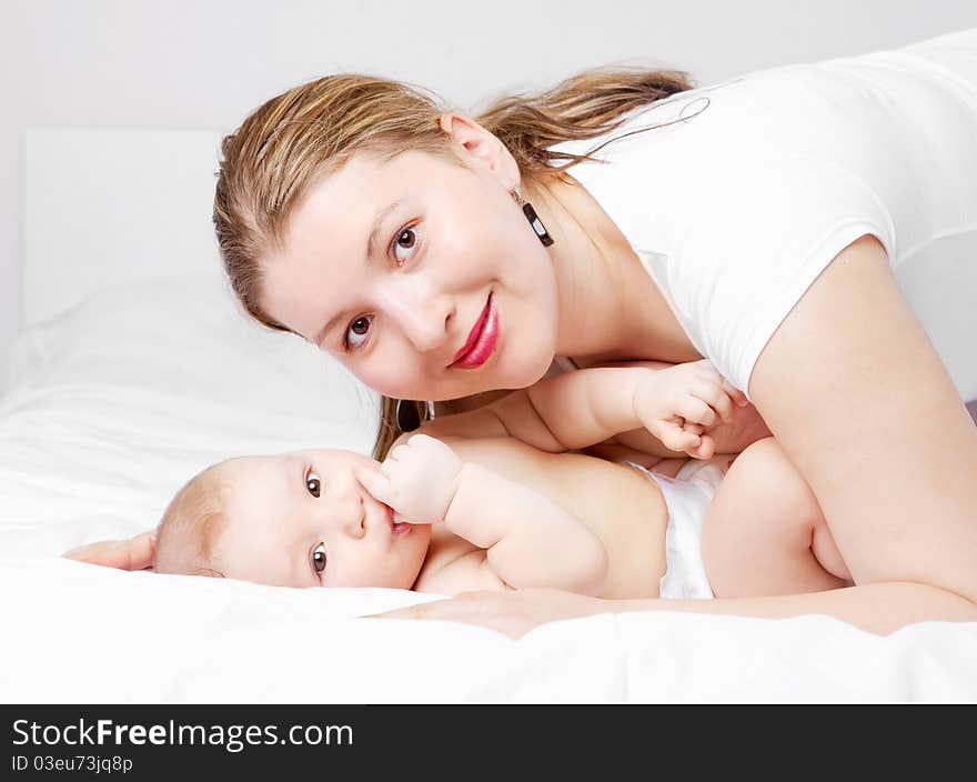 Young  mother and her six months old baby on the bed at home (focus on the baby). Young  mother and her six months old baby on the bed at home (focus on the baby)