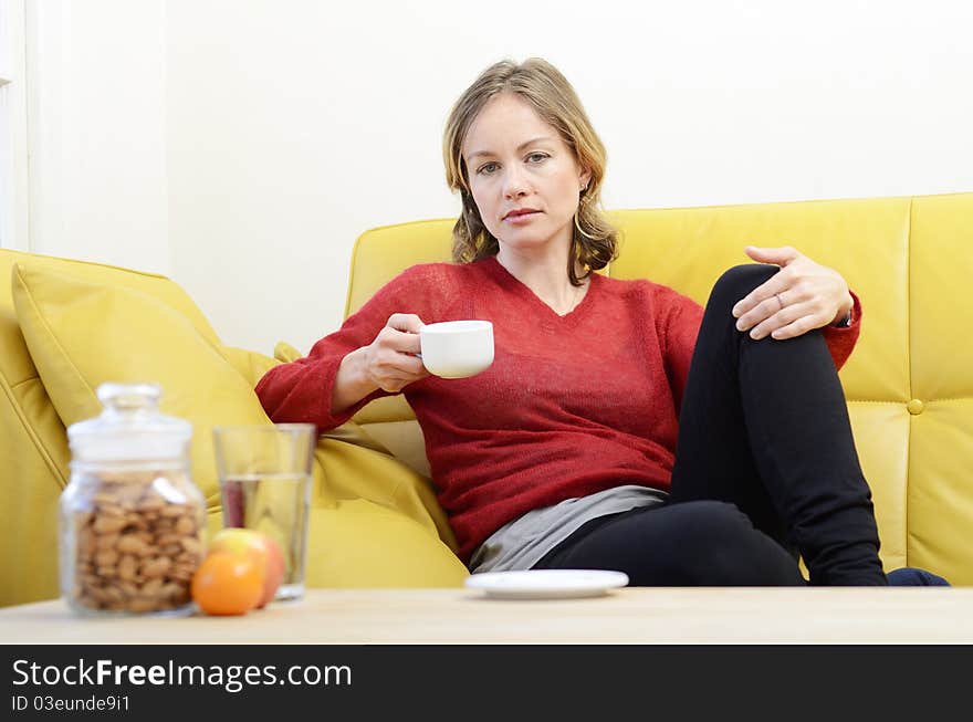 Woman sitting on a yellow leather sofa looking serious and holding a hot drink. Woman sitting on a yellow leather sofa looking serious and holding a hot drink