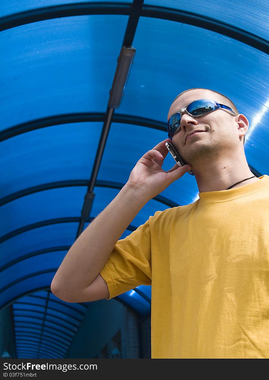 Man talking on the phone, wearing a yellow t-shirt and blue sun glasses, against blue background. Man talking on the phone, wearing a yellow t-shirt and blue sun glasses, against blue background