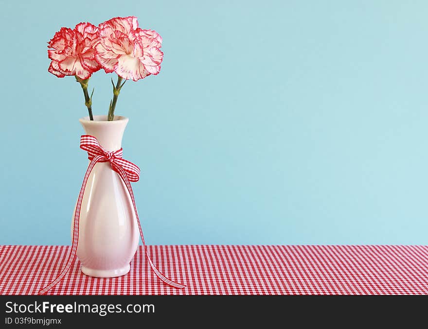 Red and white carnations in a vase on a gingham tablecloth with a light blue background