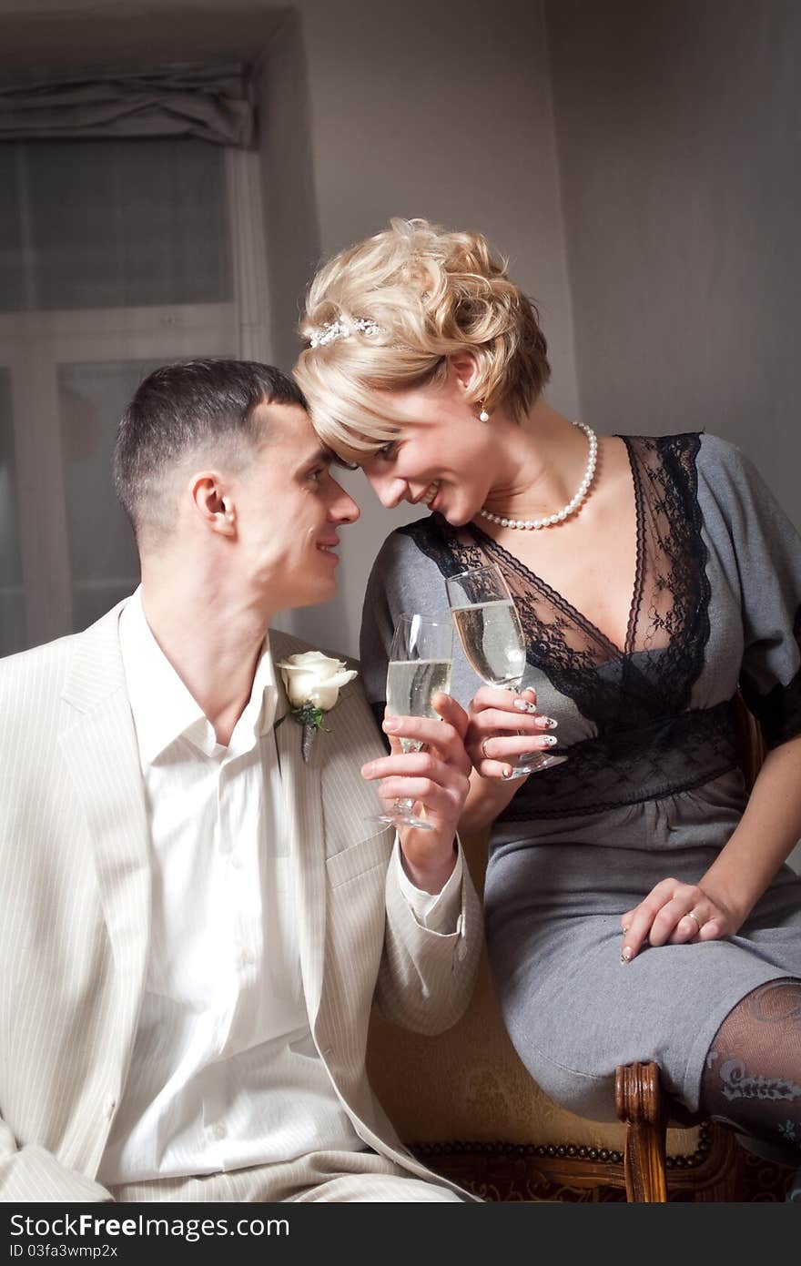 Happy bride in gray dress and groom wearing white suite in bedroom interior with glasses. Happy bride in gray dress and groom wearing white suite in bedroom interior with glasses