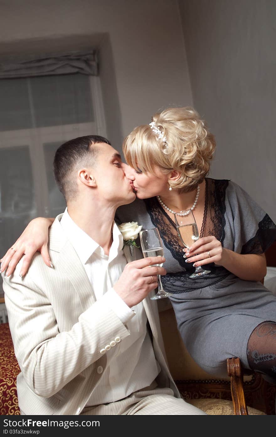 Happy bride in gray dress and groom wearing white suite are kissing in bedroom interior with glasses. Happy bride in gray dress and groom wearing white suite are kissing in bedroom interior with glasses