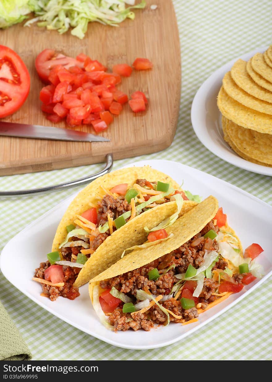 Two tacos with lettuce, tomatoes, cheese on a plate with ingredients in background