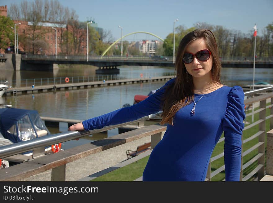 Sweet, young girl portrait. Teenage model in Poland wearing casual dress and sunglasses, smiling gently. Portrait at river side with boats as background. Sweet, young girl portrait. Teenage model in Poland wearing casual dress and sunglasses, smiling gently. Portrait at river side with boats as background.