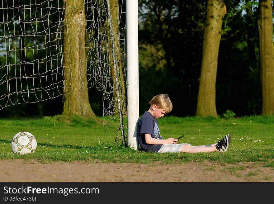 Boy playing a computer game leaning against a goal post on a football field. Boy playing a computer game leaning against a goal post on a football field